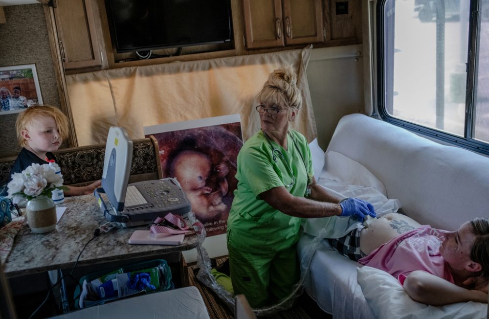 Angela Michael (67) (center), an anti-abortion activist and former nurse, performs ultrasounds in a recreational vehicle transformed into a small clinic outside the Hope Clinic for Women in Granite City, Illinois. Michael and her husband park their RV outside of the abortion clinic nearly every day to dissuade patients from seeking abortions. They offer diapers and other supplies instead. Years earlier, she convinced Ashley Miller (29) (right) not to have an abortion. Ashley is now five weeks into her 4th pregnancy, likely with twins. She had a miscarriage a few months before after being beaten by her partner, a local police officer. In the first nine months after the Supreme Court's ruling, Illinois saw an increase of 12,400 abortions from the previous year as people traveled there from states with bans, according to the Society of Family Planning. The Michaels are concerned about this surge. Granite City, Illinois, USA, 19.07.2022 



(The scene was not staged; permission was granted by Angela and Ashley, her daughter's legal guardian.)